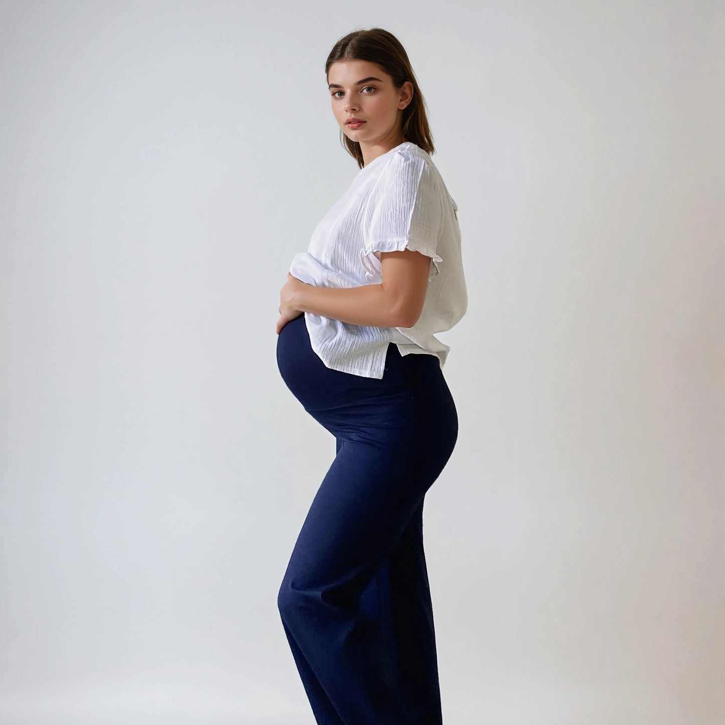 Femme enceinte portant le pantalon Cosy bleu marine, coupe droite et taille haute douce, vue de profil, sur fond studio blanc.