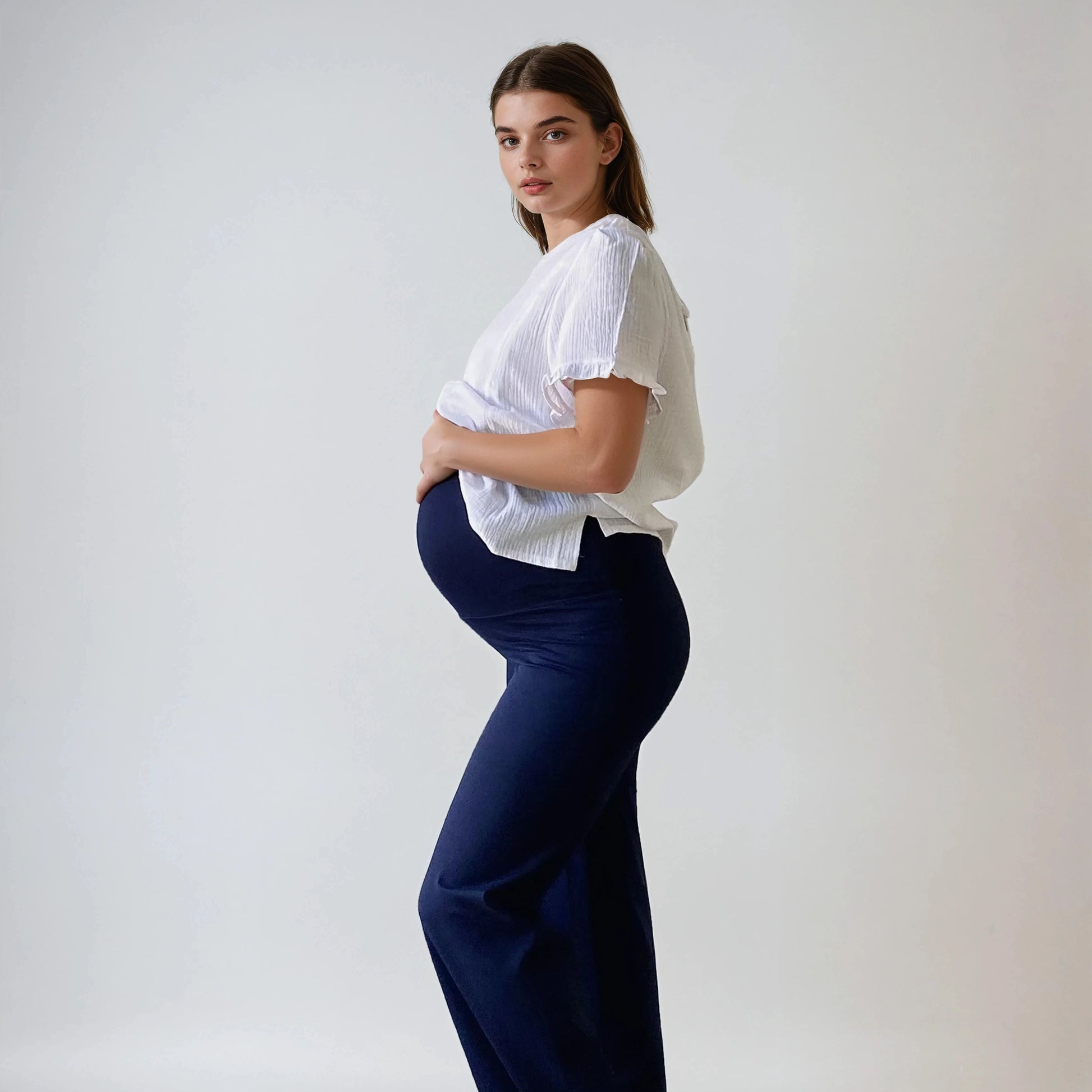 Femme enceinte portant le pantalon Cosy bleu marine, coupe droite et taille haute douce, vue de profil, sur fond studio blanc.
