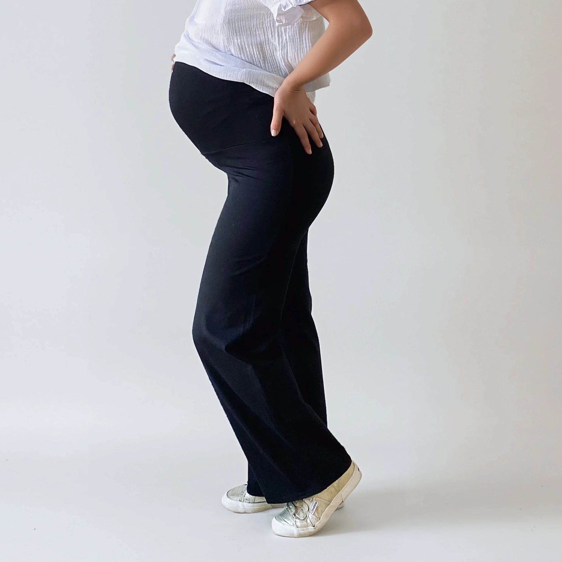Femme enceinte portant un pantalon de grossesse noir, coupe droite, taille haute confortable qui soutient le ventre, photographie sur fond studio blanc.