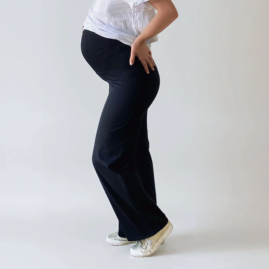Femme enceinte portant un pantalon de grossesse noir, coupe droite, taille haute confortable qui soutient le ventre, photographie sur fond studio blanc.